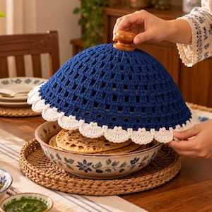May include: A dome-shaped food cover made of dark blue crocheted yarn with a white scalloped edge and a small wooden knob handle on top, positioned over a ceramic bowl containing flatbreads on a woven placemat.