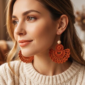 May include: A pair of burnt orange crochet earrings featuring a semi-circular fan design with intricate openwork stitching. Each earring is accented with a single round, light-colored wooden bead at the top, suspended from a simple metal hook.