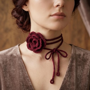 May include: A dark red crochet choker necklace featuring a large, layered rose flower accent. The necklace consists of thin, cord-like bands that wrap around the neck and tie in a bow at the front, with two dangling ends finished with small circular details.