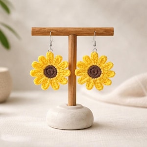 May include: A pair of handmade sunflower earrings. Each earring features a bright yellow crocheted flower with a brown center. The earrings are displayed on a wooden jewelry stand with a white base. The earrings are a charming accessory.