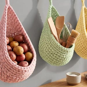 Könnte beinhalten: Drei hängende Häkelkörbe in Rosa, Grün und Gelb. Der rosa Korb enthält Kartoffeln, der grüne Küchenutensilien und der gelbe Äpfel und Zitronen. Die Körbe hängen an der Wand.