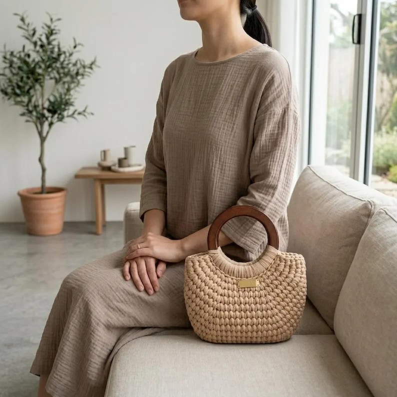 May include: A beige crocheted handbag with a rounded silhouette and a dark brown circular wooden handle. The bag features a textured, knotted stitch pattern and a small, rectangular gold-toned metal plate centered on the front exterior surface.