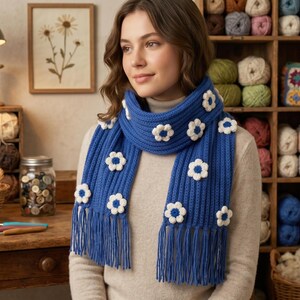 May include: A bright blue ribbed knit scarf featuring white crocheted flower appliqués with yellow centers. The scarf has long, straight fringe at both ends and is worn over a neutral-colored long-sleeved top. The design is consistent throughout.