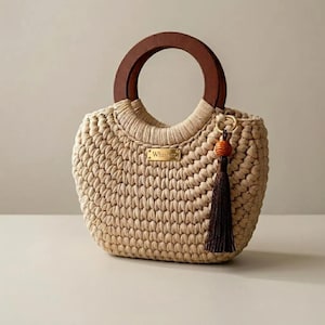 May include: A beige crocheted handbag featuring a dark brown circular wooden handle and a gold-toned metal plaque with the text Whisper. A dark brown tassel with a small wooden bead is attached to the side of the bag with a gold-toned metal ring.