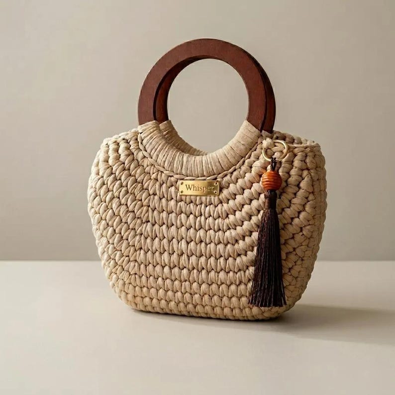 May include: A beige crocheted handbag featuring a dark brown circular wooden handle and a gold-toned metal plaque with the text Whisper. A dark brown tassel with a small wooden bead is attached to the side of the bag with a gold-toned metal ring.