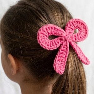 May include: A pink crocheted bow hair clip is attached to the back of a person's head. The bow is made of a thick yarn and is a bright, solid pink color. The person's hair is brown and pulled back in a ponytail.