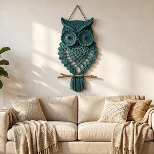 May include: A dark teal macrame owl wall hanging featuring intricate knotted patterns, large circular eyes with dark centers, and a fringe tail suspended from a horizontal wooden branch with a thin cord for mounting on a wall surface.