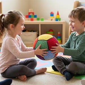 Puede incluir: Niños jugando con una pelota de ganchillo multicolor. La pelota presenta secciones rojas, amarillas, verdes y azules. El fondo muestra estanterías con juguetes, creando un ambiente lúdico.