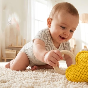 Op de afbeelding: Een lachende baby in een gestreepte romper speelt met een houten ring en een geel hartvormig speeltje op een crèmekleurig tapijt. De kinderkamer heeft een commode met pastelkleurige lades en ingelijste kunst.