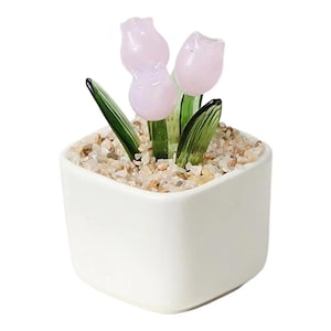 May include: A small, square white ceramic pot with three pink glass tulip flowers and green glass leaves. The pot is filled with small, light-colored pebbles. The tulips have a delicate, translucent appearance.
