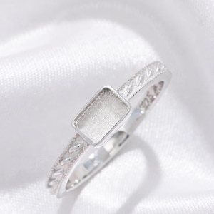 May include: A silver ring featuring a rectangular bezel setting for a stone. The band has a textured, rope-like pattern along the sides and is stamped with 925 on the inner surface. The metal has a polished finish and a bright, reflective appearance.