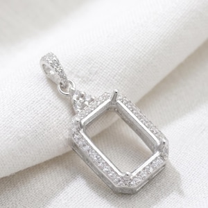 Puede incluir: Un colgante rectangular de tono plateado con un centro hueco y un halo de pequeñas piedras claras y brillantes. El colgante incluye una reasa decorativa en la parte superior, también incrustada con pequeñas piedras claras.