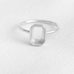 Könnte beinhalten: Ein silberfarbener Ring mit einer dünnen, glatten Ringschiene und einer mittigen, leeren rechteckigen Fassung mit abgeschrägten Kanten. Die Fassung ist für die Aufnahme eines Steins oder Einsatzes vorgesehen und hat ein klares Metallfinish.