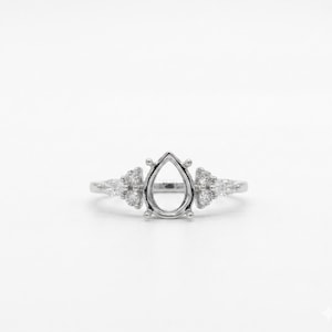May include: A silver ring with a pear-shaped setting, flanked by clusters of small, round and pear-shaped stones. The ring is set against a white background, highlighting its intricate design and sparkle. A delicate piece of jewelry.
