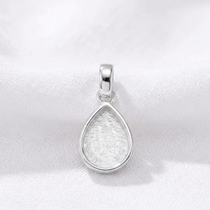 May include: A silver-toned pendant featuring a teardrop-shaped bezel setting with a textured, matte-finish interior surface. The pendant includes a polished, smooth bail at the top for attachment to a chain, showcasing a clean and simple design.