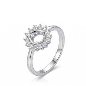May include: A silver ring with a halo design. The ring features a central setting surrounded by a cluster of small, sparkling stones, creating a sunburst effect. The band is smooth and polished, with a simple, elegant design.