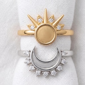 May include: A gold ring featuring a central circular disc surrounded by radiating spikes and small clear stones, paired with a silver ring featuring a crescent moon shape adorned with a row of small clear stones along the outer edge.