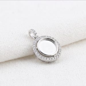 May include: A circular silver-toned pendant featuring a central recessed circular setting designed for an inlay. The outer edge of the pendant is framed by a halo of small, clear, sparkling stones, and it includes a small bail at the top.