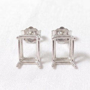 May include: A pair of silver-colored stud earrings. Each earring features a rectangular frame design with four small feet. The earrings are set against a white background.