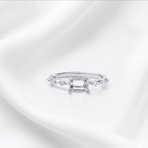 May include: A silver engagement ring with a square center setting and small accent stones along the band. The ring is set against a white background, highlighting its design and details. The ring is a classic style.
