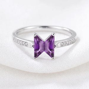 May include: A silver-toned ring featuring a central purple gemstone cut into a trapezoidal shape that resembles a vest icon. The band is polished and includes a row of small, clear, round-cut stones embedded along each side of the shank.