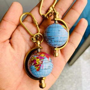 May include: Two miniature globe keychains with gold-colored metal hardware. The globes are painted with a detailed map of the world, featuring various countries and oceans. The keychains have a swivel design and a clasp for attaching to keys or bags.