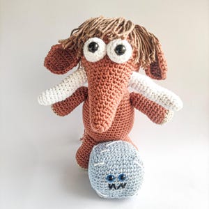 Puede incluir: Una muñeca de elefante de ganchillo hecha a mano en tonos marrón, blanco y beige, con grandes ojos saltones y un compañero azul y redondo con forma de monstruo. El elefante tiene pelo largo y con flecos y colmillos blancos. El monstruo tiene ojos azules y boca cosida.