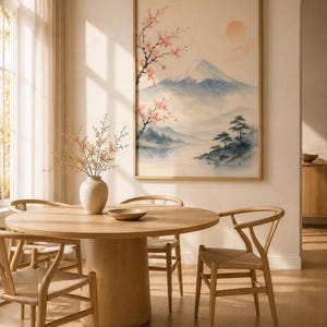 A watercolor print of Mount Fuji with cherry blossoms and a red sun, in a light wood frame, hanging above a round wooden dining table with four chairs.