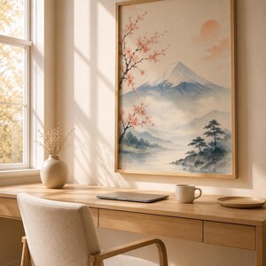 A watercolor print of Mount Fuji with cherry blossoms and a red sun, in a light wood frame, hanging above a wooden desk with a laptop, mug, and a vase with dried branches.