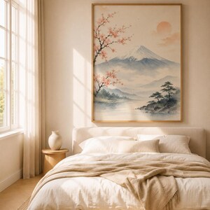A watercolor print of Mount Fuji with cherry blossoms and a red sun, in a light wood frame, hanging above a bed with cream bedding and a white vase on a side table.
