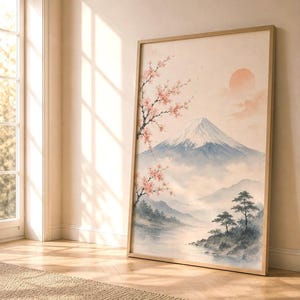 A watercolor print of Mount Fuji with cherry blossoms and a red sun, in a light wood frame, leaning against a cream wall next to a window.