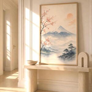 A watercolor print of Mount Fuji with cherry blossoms and a red sun, in a light wood frame, hanging above a light-colored console table with a bowl and a U-shaped sculpture.