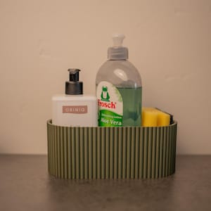 Könnte beinhalten: Ein grüner, gerippter Behälter enthält einen weißen Seifenspender, eine transparente Flasche Spülmittel mit grünem Frosch-Etikett und einen gelben Schwamm. Der Behälter steht auf einer grauen Oberfläche.