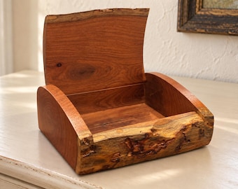 Puede incluir: Una caja de madera con la tapa abierta, que presenta un interior liso de color marrón rojizo y un borde natural de textura rugosa en el panel frontal. Los lados y la tapa están hechos de madera pulida, mostrando un veteado distintivo.