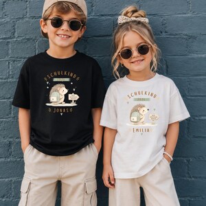 May include: A black t-shirt and a white t-shirt, each featuring a graphic of a hedgehog with a backpack, a signpost, and the text SCHULKIND 2026. The black shirt includes the name JONAS, while the white shirt includes the name EMILIA.