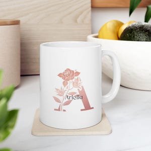 May include: A white ceramic mug featuring a large, dusty rose-colored letter A decorated with a floral rose illustration. The name Ariella is printed in black, sans-serif text across the center of the letter. The mug has a handle on the right side.