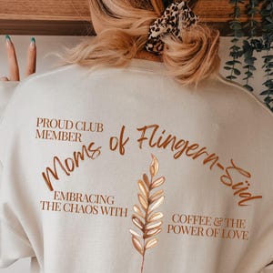 Puede incluir: Una sudadera de color crema con el texto "Moms of Flingern-Süd" en escritura color cobre. La parte trasera de la sudadera también presenta las frases "Proud Club Member", "Embracing the Chaos With" y "Coffee & the Power of Love."