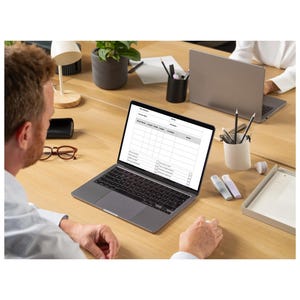 Pu&ograve; includere: Un computer portatile con un foglio di calcolo sullo schermo. Il portatile &egrave; su una scrivania di legno chiaro, con un altro portatile visibile sullo sfondo. Altri oggetti sulla scrivania includono un portapenne, matite e una piccola pianta in vaso.