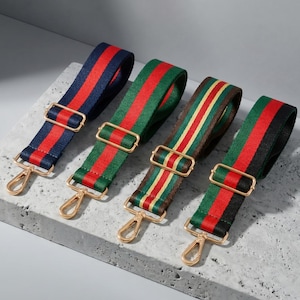 Pode incluir: Quatro alças de bolsa listradas coloridas com ferragens de metal dourado. As alças apresentam listras vermelhas e azuis, vermelhas e verdes, marrons e amarelas e vermelhas e pretas. Cada alça tem uma fivela retangular e um mosquetão.