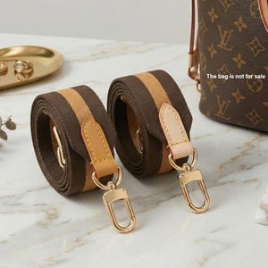 May include: Two rolled brown and tan striped purse straps with gold-tone hardware and leather accents. The straps are displayed on a marble surface. The bag is not for sale.
