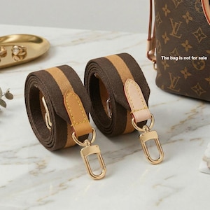 May include: Two rolled bag straps made of brown and tan woven fabric. Each strap features a light tan leather tab with yellow stitching and a gold-toned metal lobster clasp. The straps are positioned next to a brown patterned bag.