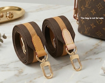 May include: Two rolled bag straps made of brown and tan woven fabric. Each strap features a light tan leather tab with yellow stitching and a gold-toned metal lobster clasp. The straps are positioned next to a brown patterned bag.