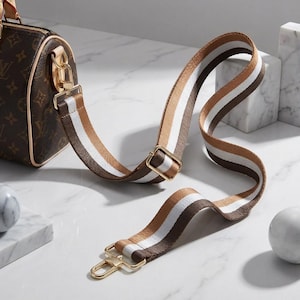Peut inclure: Un sac &agrave; main marron avec une bandouli&egrave;re ray&eacute;e marron, blanche et beige. La bandouli&egrave;re est fix&eacute;e avec des attaches dor&eacute;es. Le sac pr&eacute;sente un motif r&eacute;p&eacute;t&eacute;. La bandouli&egrave;re est con&ccedil;ue pour &ecirc;tre port&eacute;e &agrave; l'&eacute;paule.