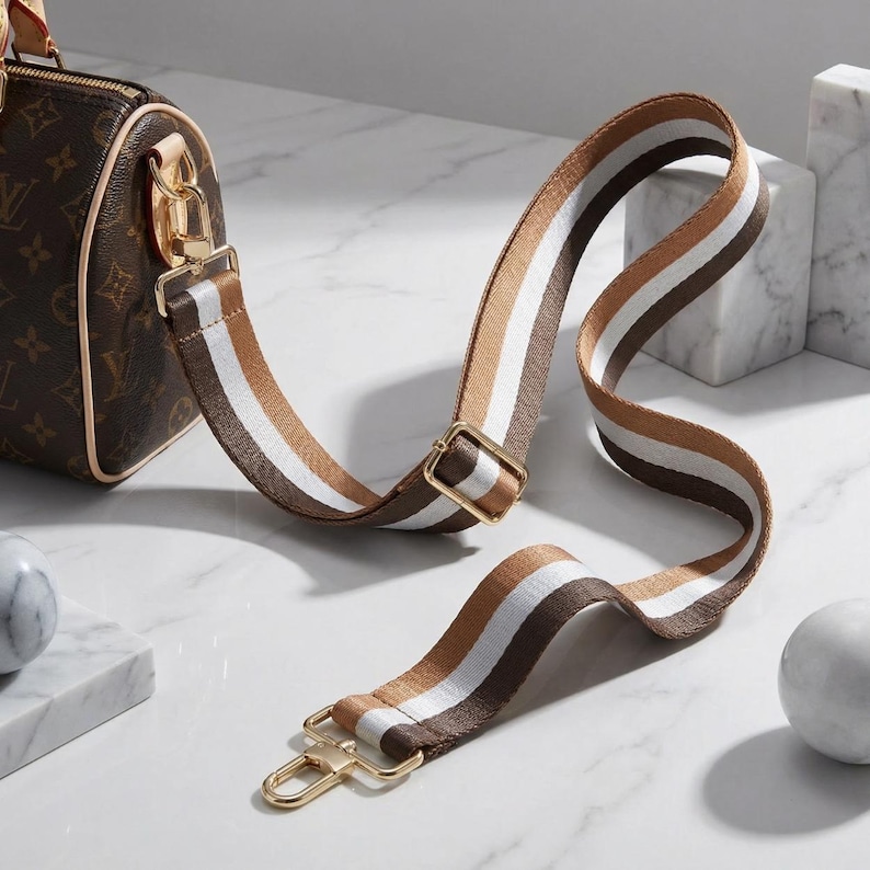 Peut inclure: Un sac &agrave; main marron avec une bandouli&egrave;re ray&eacute;e marron, blanche et beige. La bandouli&egrave;re est fix&eacute;e avec des attaches dor&eacute;es. Le sac pr&eacute;sente un motif r&eacute;p&eacute;t&eacute;. La bandouli&egrave;re est con&ccedil;ue pour &ecirc;tre port&eacute;e &agrave; l'&eacute;paule.