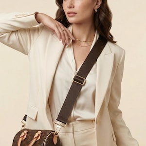 Peut inclure: Un sac &agrave; main marron avec une bandouli&egrave;re assortie. La bandouli&egrave;re est fix&eacute;e au sac avec une quincaillerie dor&eacute;e. Le sac est marron avec un motif r&eacute;p&eacute;titif. La femme porte un blazer cr&egrave;me et une chemisier blanc.