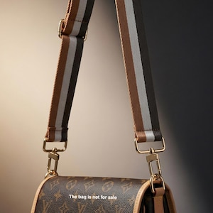 Peut inclure: Gros plan sur un sac &agrave; main marron avec un motif et une bandouli&egrave;re ray&eacute;e. La bandouli&egrave;re pr&eacute;sente des rayures marron, grises et noires. Le sac a une quincaillerie dor&eacute;e. Le texte sur le sac indique : "Le sac n'est pas &agrave; vendre."