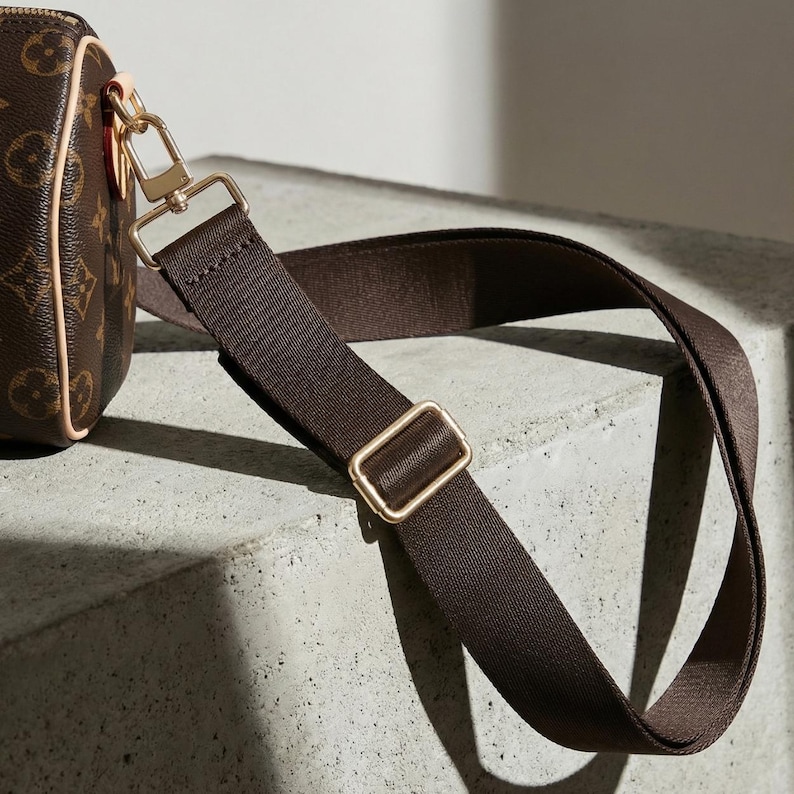 Peut inclure: Gros plan sur un sac bandouli&egrave;re marron avec une bandouli&egrave;re r&eacute;glable assortie. Le sac est dot&eacute; d'un fermoir et d'une boucle dor&eacute;s. L'ext&eacute;rieur du sac pr&eacute;sente un motif r&eacute;p&eacute;titif. La bandouli&egrave;re est en mati&egrave;re tiss&eacute;e durable.