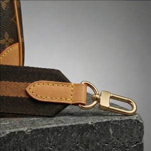 Op de afbeelding: Close-up van een bruine en tan handtasriem met een goudkleurige sluiting. De riem is gemaakt van een donkerbruine stof met een tan streep en een tan leren eindstuk. De sluiting heeft een glanzende goudkleur.