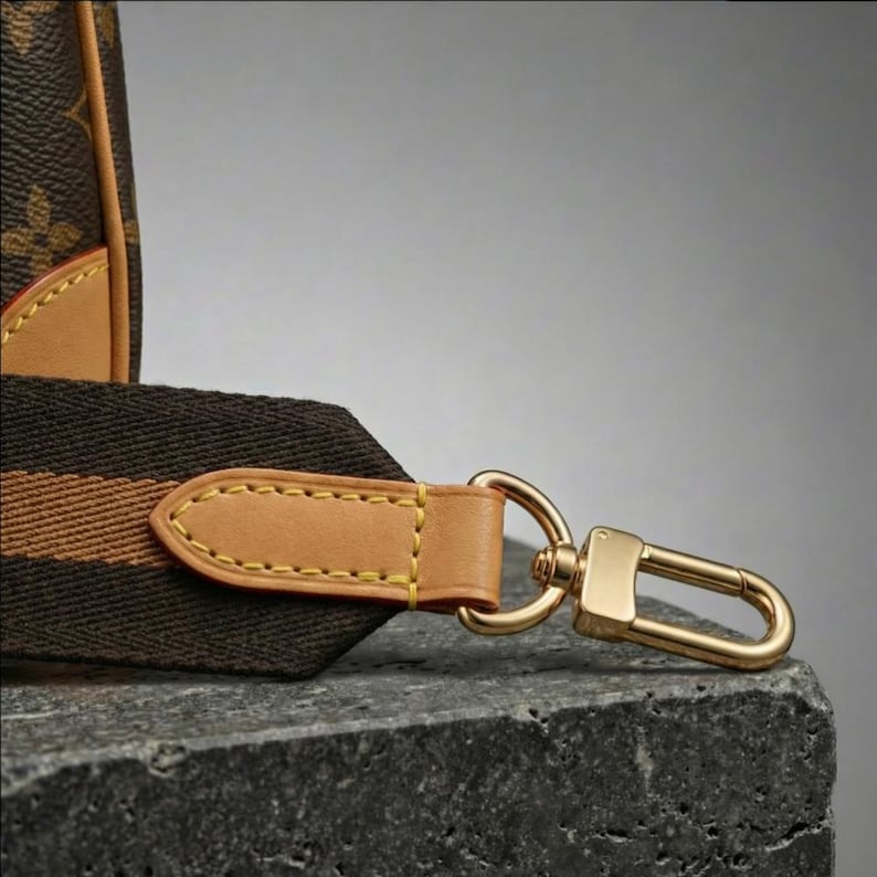 Op de afbeelding: Close-up van een bruine en tan handtasriem met een goudkleurige sluiting. De riem is gemaakt van een donkerbruine stof met een tan streep en een tan leren eindstuk. De sluiting heeft een glanzende goudkleur.