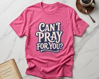May include: A bright pink short-sleeved t-shirt featuring the text "CAN I PRAY FOR YOU?" in a bold, dark blue and cream-coloured stylised font. The shirt has a crew neckline and is displayed flat, showcasing the graphic design centred on the chest.